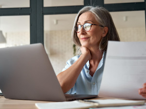 Digitalisierung: Ältere Dame sitzt lächelnd mit einem Papier in der Hand vor ihrem Laptop