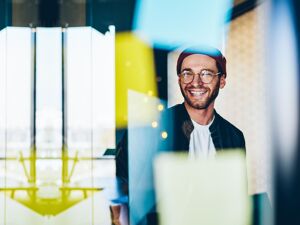 Fröhlicher Mann steht lächelnd vor einer Glaswand mit Post-ITs